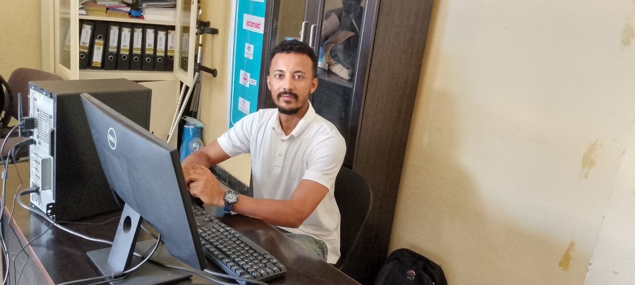 Shewit Enquebaher, Program Director of TDOU — a young man in a white polo shirt seated at his desk in front of a computer.
