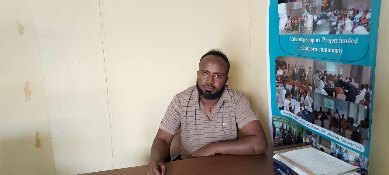 Tekeste Desta, Monitoring, Evaluation and Learning Officer at TDOU — a man with a short beard in a beige striped polo, seated at a table in front of a TDOU program banner.