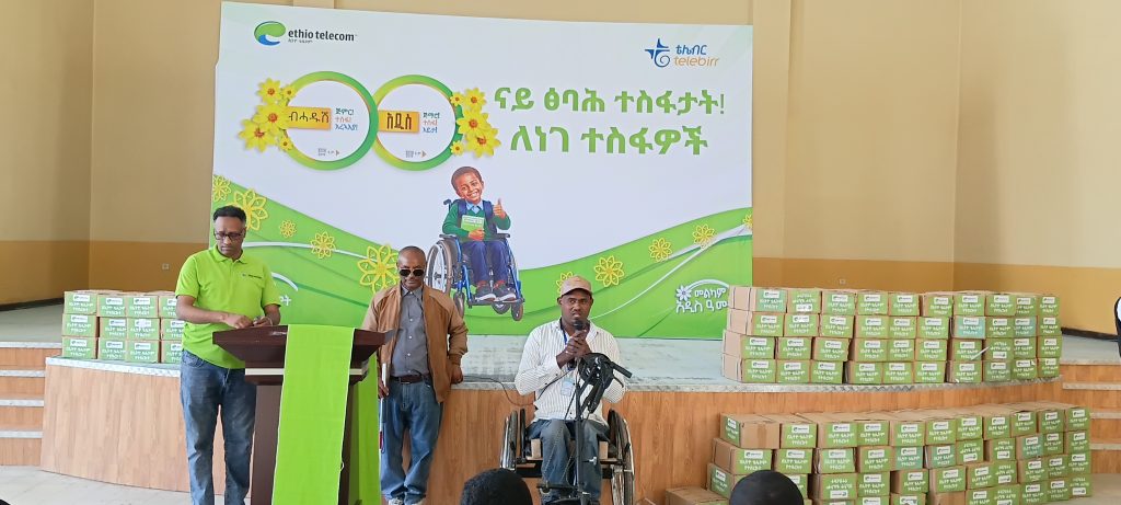 A man seated in a hand-cycle wheelchair speaks into a microphone on a stage, with two men standing nearby, one at a podium, and stacks of boxed exercise books and an Ethio telecom banner behind them