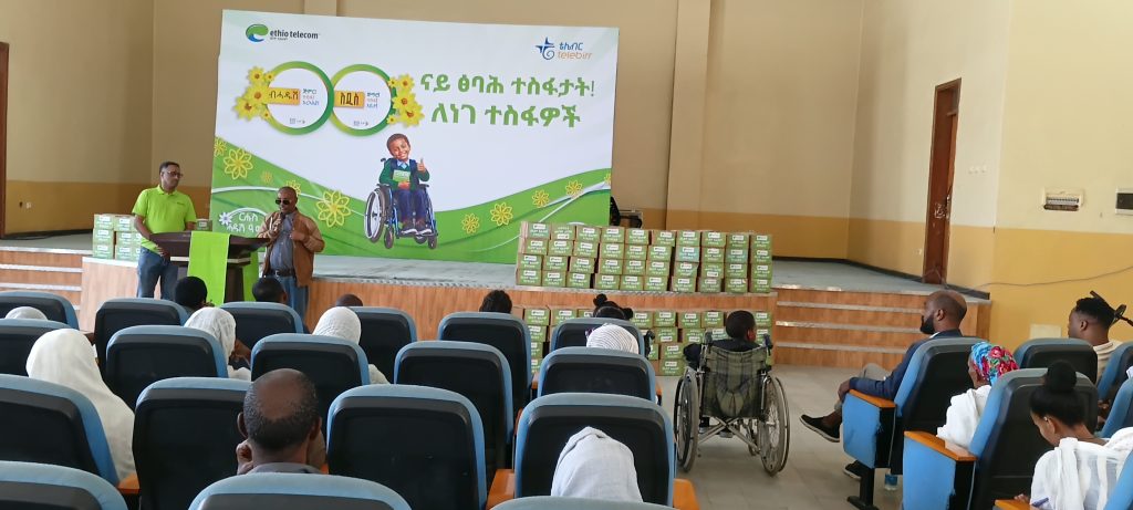 A man in a brown jacket and dark glasses speaks into a microphone on stage beside a podium, while an audience sits in blue chairs in the foreground, including a person in a wheelchair, with stacks of boxed exercise books and an Ethio telecom banner behind.