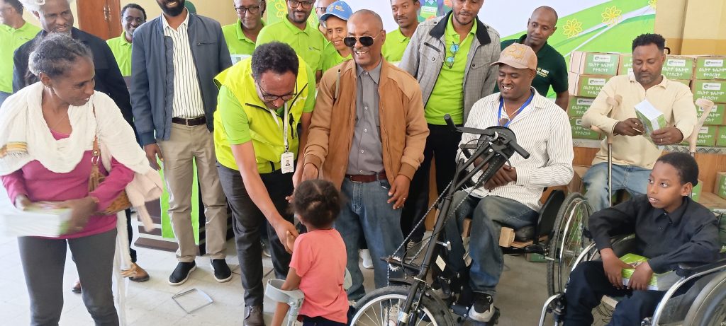 An Ethio telecom representative in a green vest bends down to greet a young child using a forearm crutch, surrounded by smiling attendees, some in wheelchairs, with boxes of exercise books stacked behind them